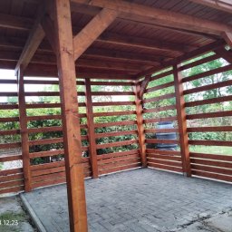 robert nowacki - Drewniana konstrukcja z zadaszeniem i ażurowymi ścianami z poziomych desek, posadowiona na kostce brukowej, z widocznym zbiornikiem na deszczówkę w tle.