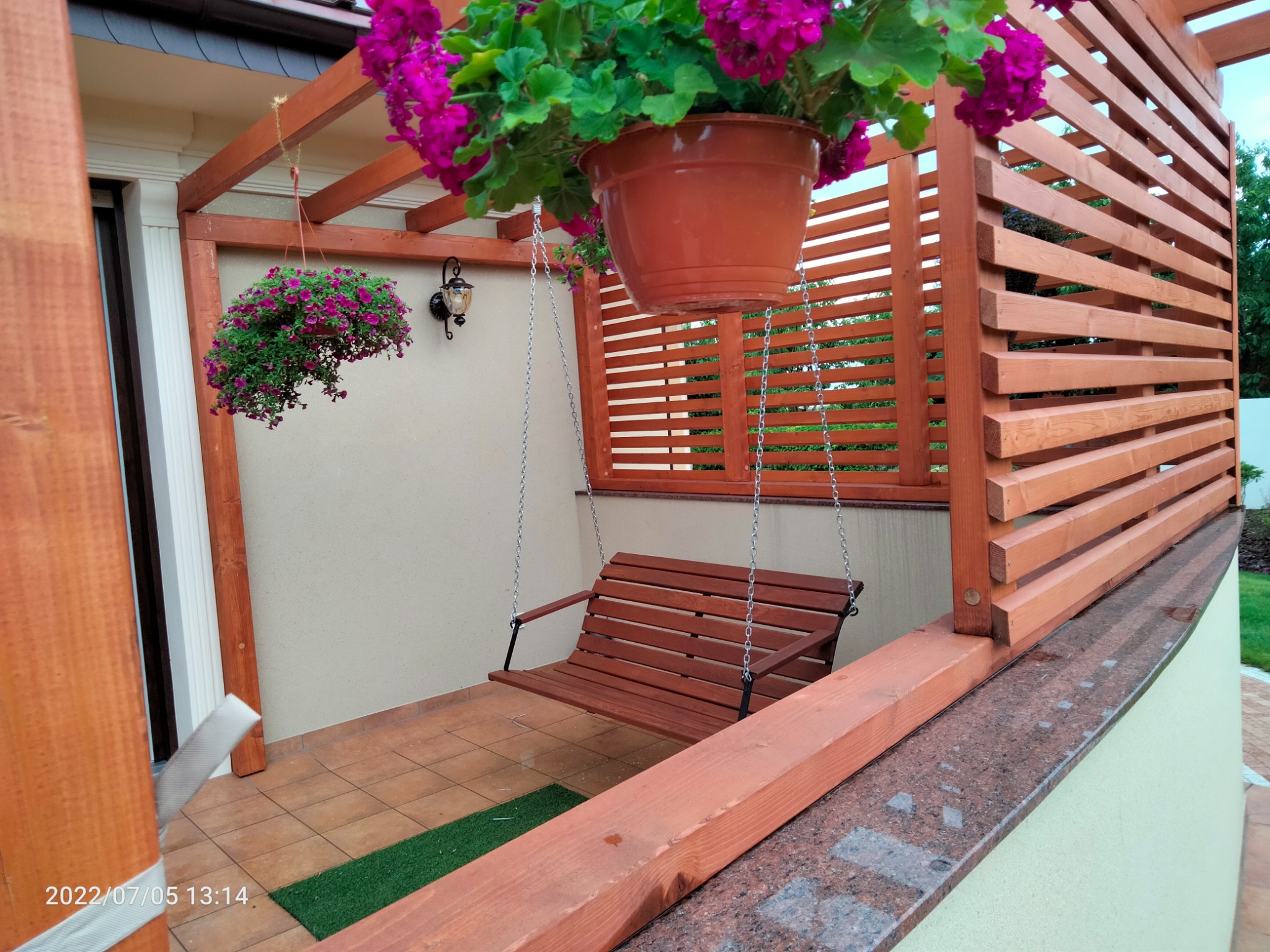 Zadaszony taras z drewnianą konstrukcją, wiszącą huśtawką na łańcuchach i doniczkami z kwiatami.