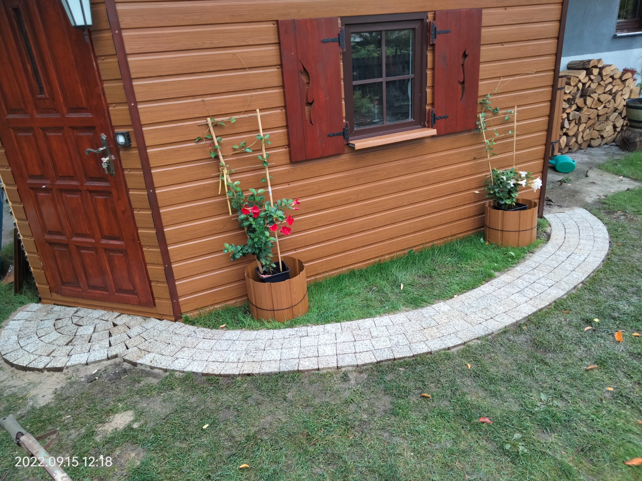 Ułożony z kostki granitowej chodnik prowadzący do drewnianej altany z brązowymi okiennicami, z donicami z kwiatami po bokach.