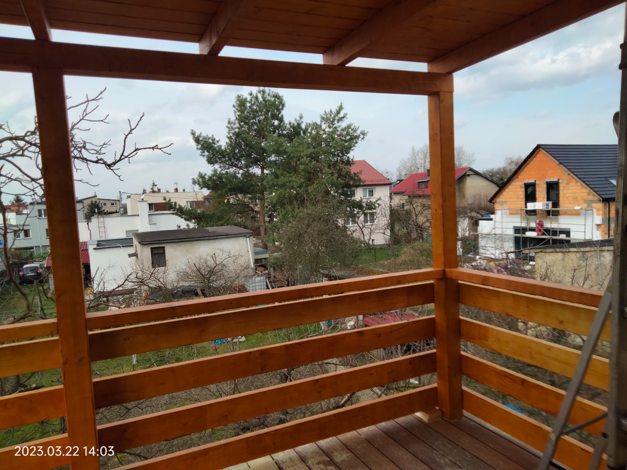 Drewniany taras z balustradą w trakcie budowy, widok na okoliczne domy i drzewa, drabina oparta o konstrukcję.