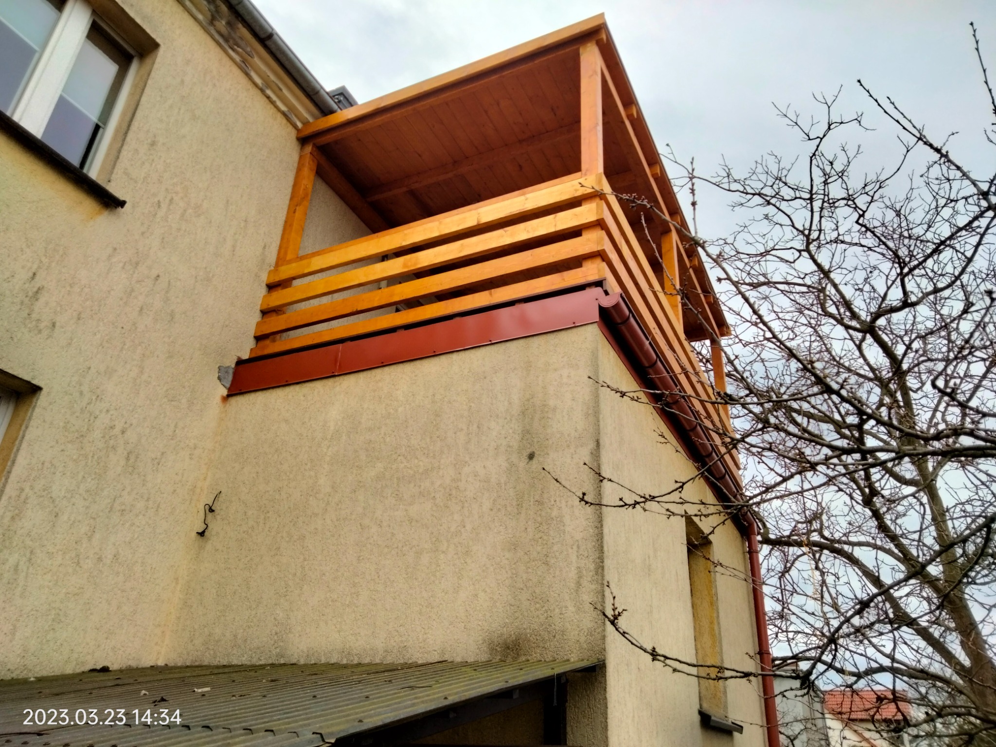 Drewniana konstrukcja zadaszonego tarasu na piętrze budynku z beżową elewacją, widoczna z dołu pod kątem, z nagimi gałęziami drzewa w tle.