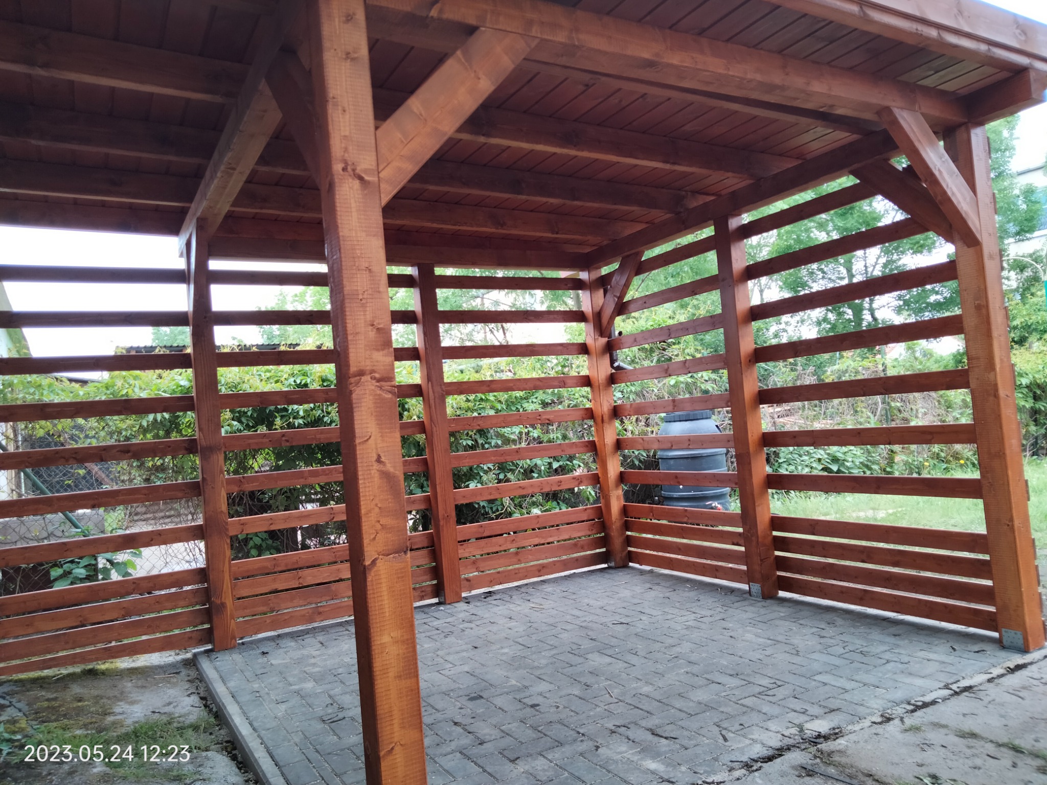 Drewniana konstrukcja z zadaszeniem i ażurowymi ścianami z poziomych desek, posadowiona na kostce brukowej, z widocznym zbiornikiem na deszczówkę w tle.