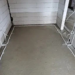 Surowy stan: Białe przewody elektryczne ułożone na świeżym, szarym betonie w pomieszczeniu z białymi ścianami z bloczków betonowych.