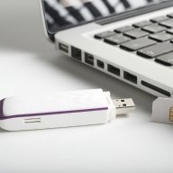 Biały modem USB z fioletowym akcentem, karta SIM i fragment laptopa na białym tle. Sprzęt do łączenia z internetem.