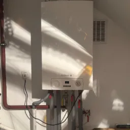 Biały piec gazowy De Dietrich zamontowany na ścianie z widocznymi rurami, zaworami i elementami instalacji. Ściana oświetlona padającym światłem.