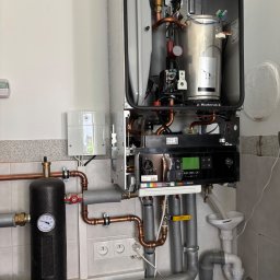 PrimeMont Damian Cebula - Otwarty, srebrny piec gazowy Buderus z widocznymi miedzianymi rurami, podłączony do instalacji z czarnym zbiornikiem ciśnieniowym. W tle białe płytki.