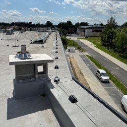 PrimeMont Damian Cebula - Instalacja odgromowa na płaskim dachu budynku z kominem i panelami słonecznymi w tle. Widok z góry, słoneczny dzień.