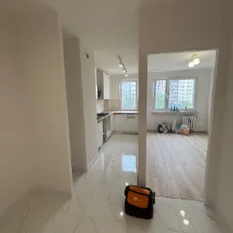 Jasne wnętrze po remoncie: białe ściany, marmurowe płytki podłogowe, widok na kuchnię i salon. Torba narzędziowa w rogu kadru.