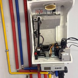 PrimeMont Damian Cebula - Otwarty piec gazowy z kolorowymi rurami (żółtą, czerwoną, niebieską) przy ścianie. Widoczne elementy wewnętrzne pieca i podłączenia.