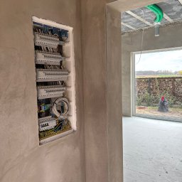 PrimeMont Damian Cebula - Rozdzielnica elektryczna w surowym wnętrzu domu, widoczne przewody i zabezpieczenia. Przez okno w tle widać ułożone drewno i krajobraz.