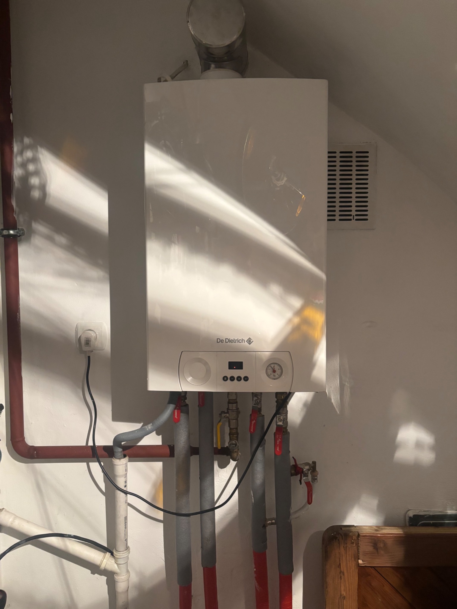 Biały piec gazowy De Dietrich zamontowany na ścianie z widocznymi rurami, zaworami i elementami instalacji. Ściana oświetlona padającym światłem.