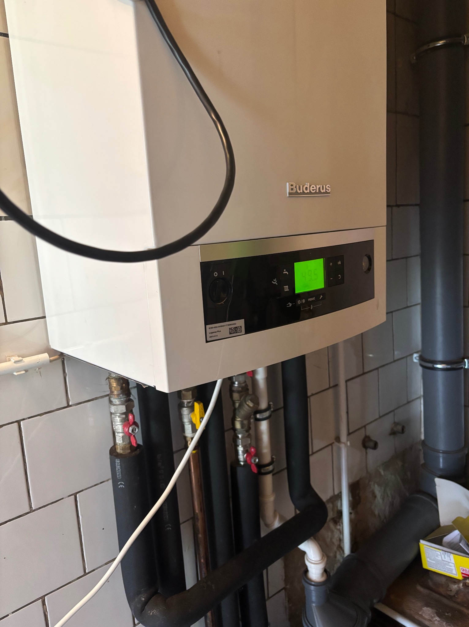 Biały piec gazowy Buderus z podłączonymi rurami, zamontowany na ścianie z białymi płytkami. Widoczne zawory i wyświetlacz z zielonymi cyframi.