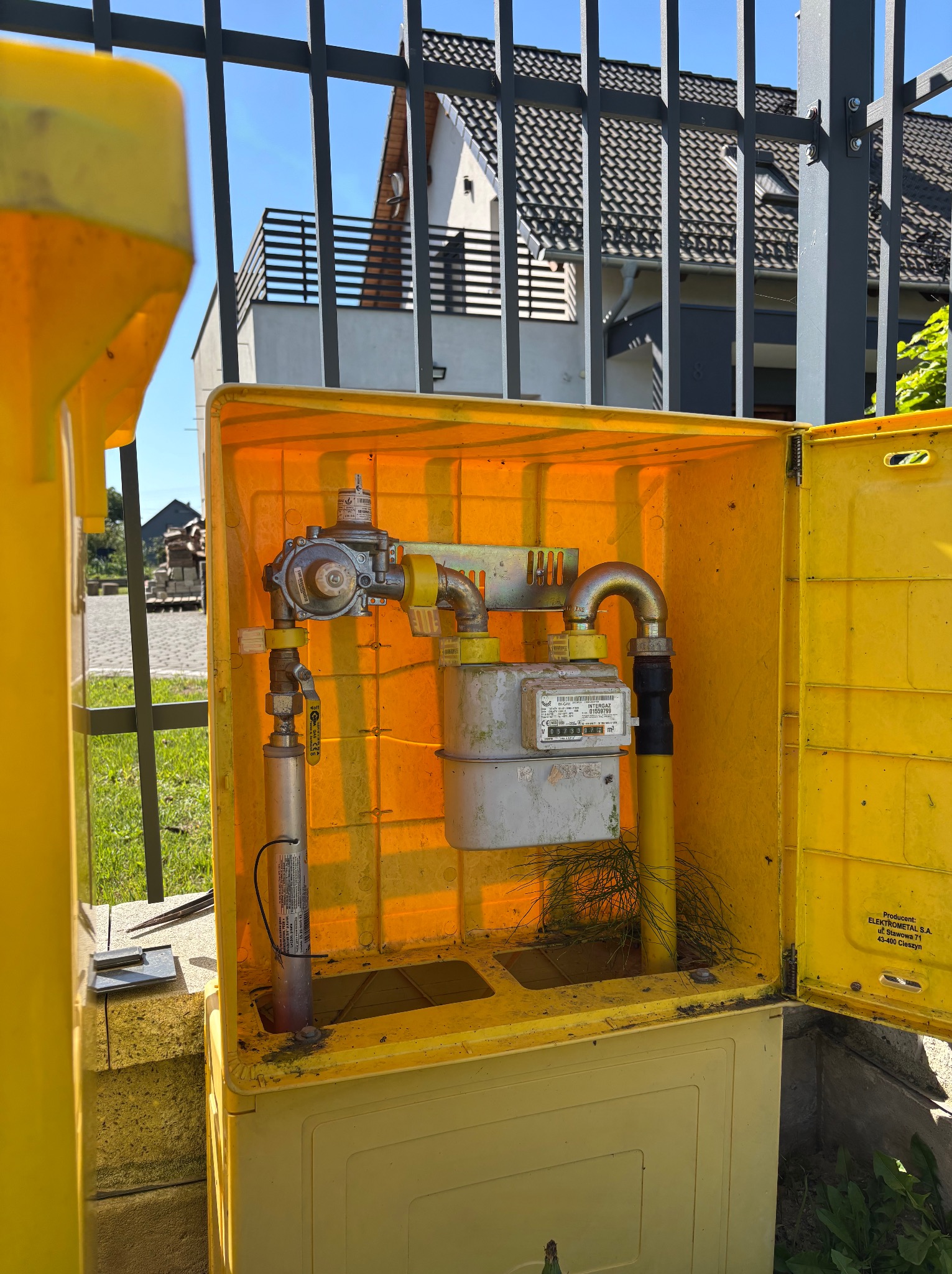 Żółta skrzynka gazowa z licznikiem na tle ogrodzenia i domu. Widoczne elementy instalacji gazowej, rury i zawory. Skrzynka otwarta, z widocznymi zabrudzeniami wewnątrz.