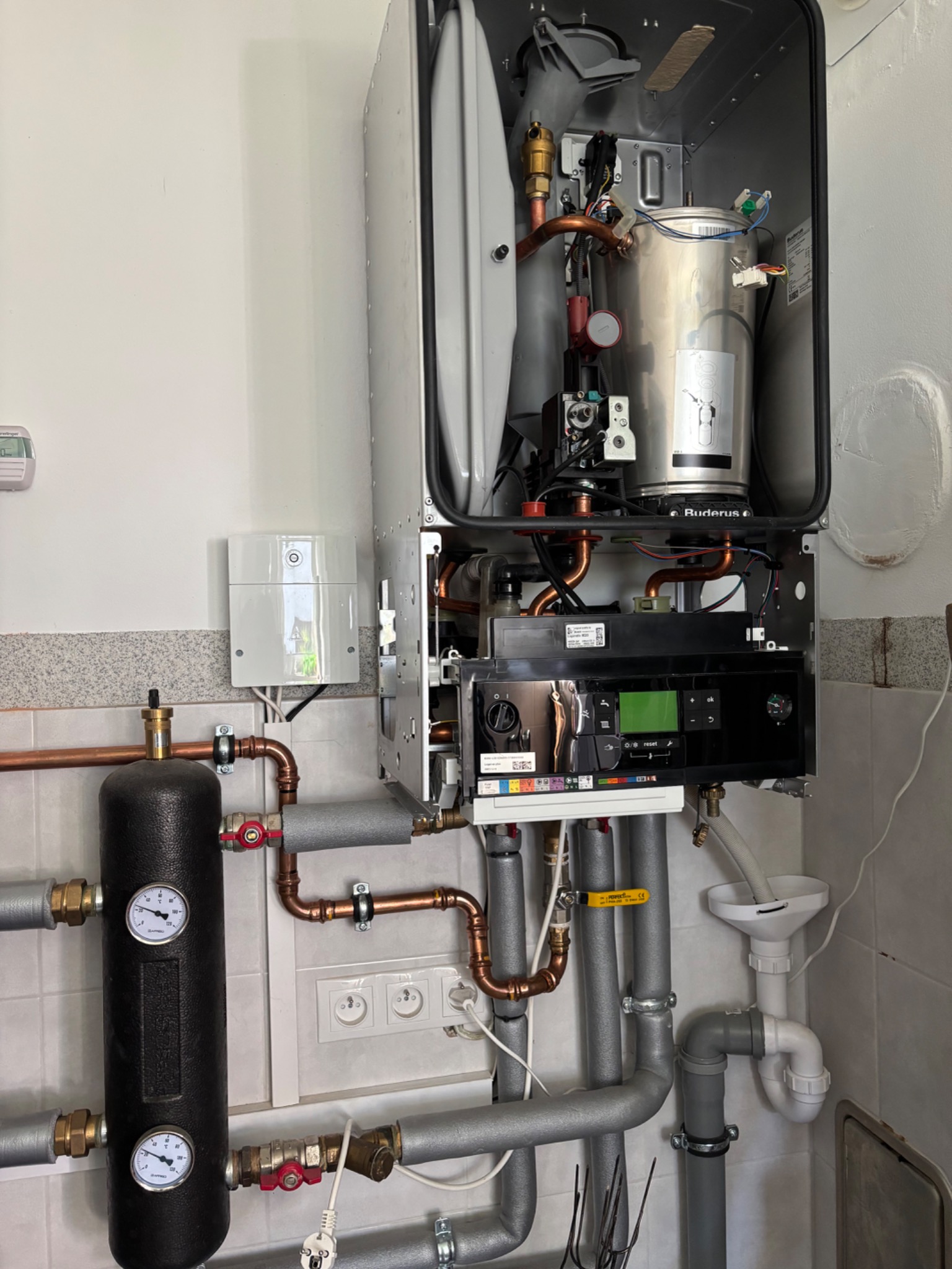 Otwarty, srebrny piec gazowy Buderus z widocznymi miedzianymi rurami, podłączony do instalacji z czarnym zbiornikiem ciśnieniowym. W tle białe płytki.