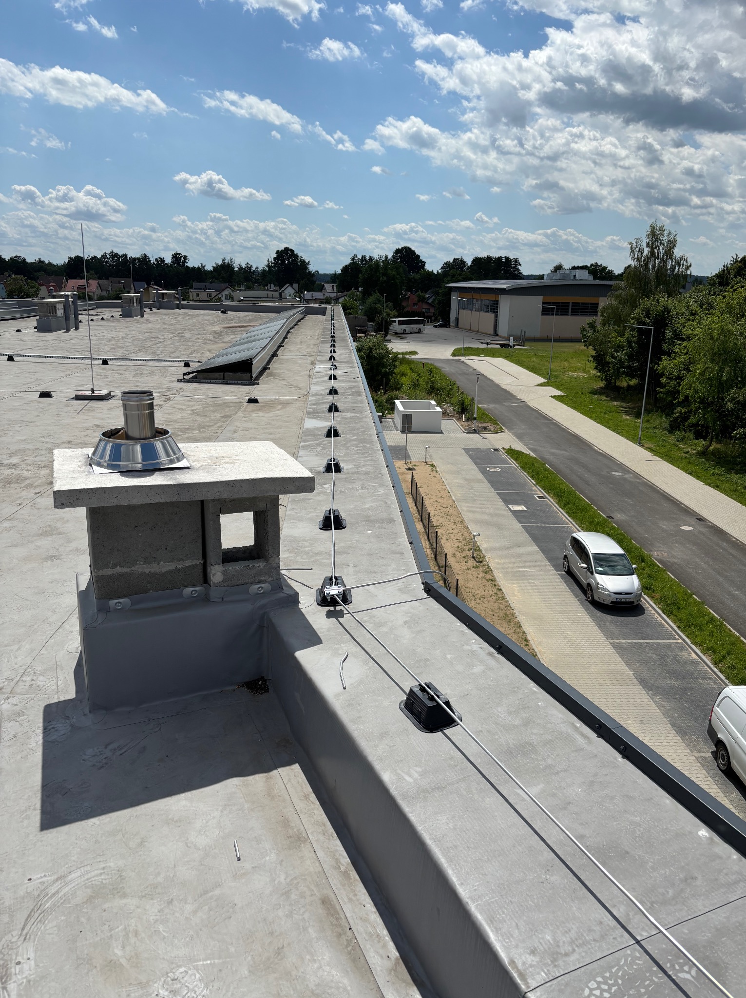 Instalacja odgromowa na płaskim dachu budynku z kominem i panelami słonecznymi w tle. Widok z góry, słoneczny dzień.