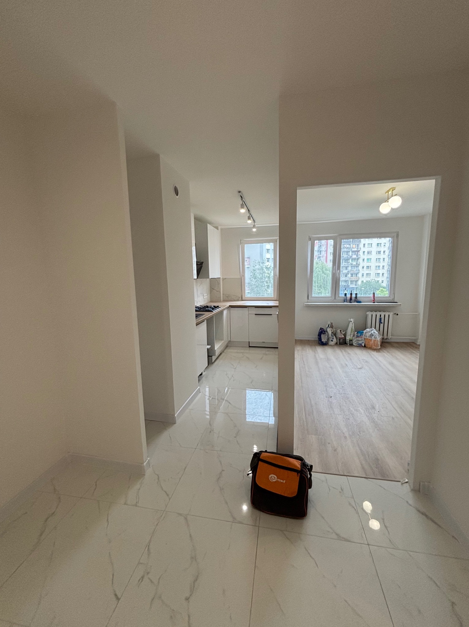 Jasne wnętrze po remoncie: białe ściany, marmurowe płytki podłogowe, widok na kuchnię i salon. Torba narzędziowa w rogu kadru.