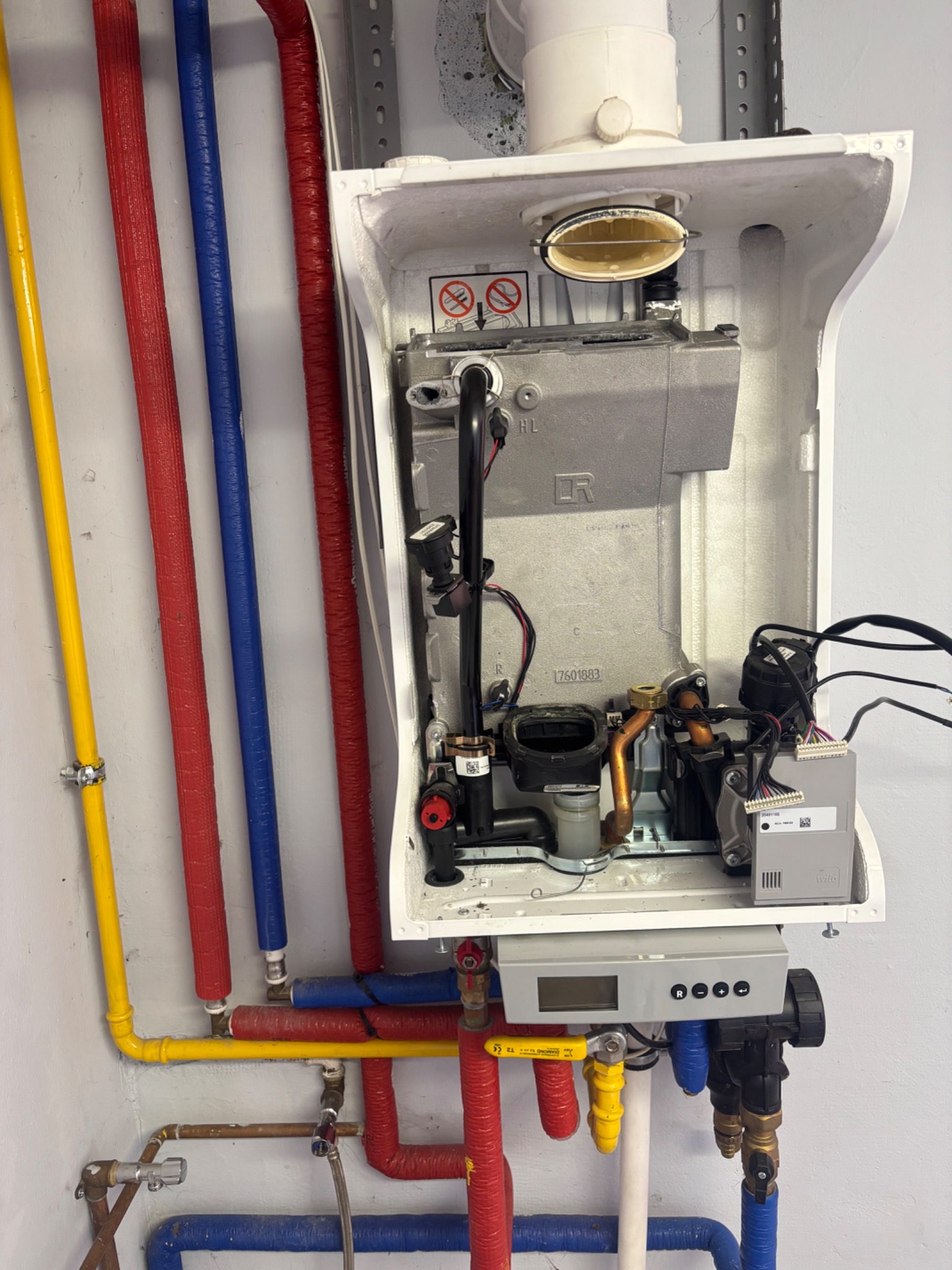 Otwarty piec gazowy z kolorowymi rurami (żółtą, czerwoną, niebieską) przy ścianie. Widoczne elementy wewnętrzne pieca i podłączenia.