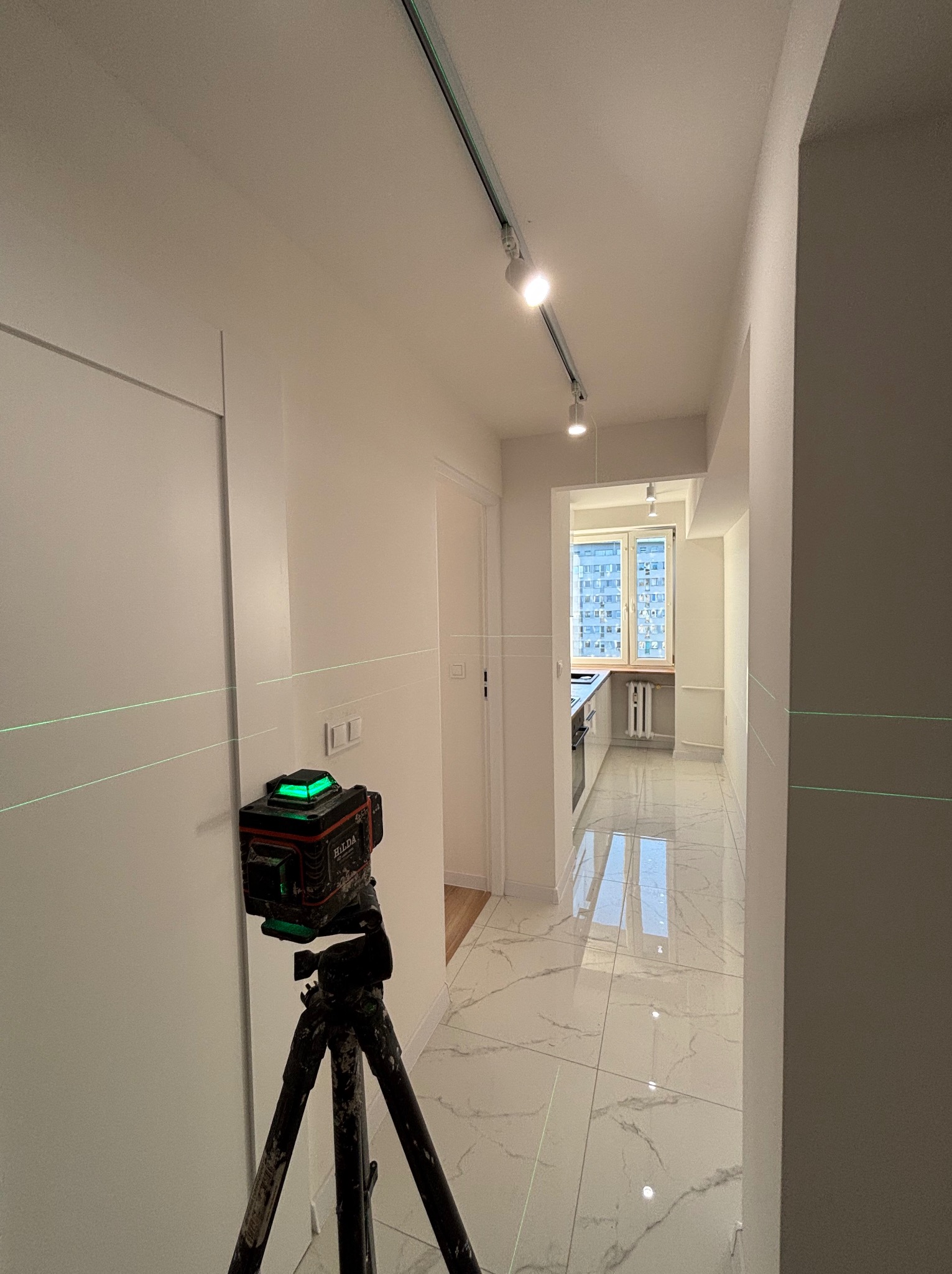 Laser krzyżowy Hilda na statywie, emitujący zielone linie na ścianach korytarza z oświetleniem szynowym. W tle widoczna kuchnia z oknem i marmurową podłogą.