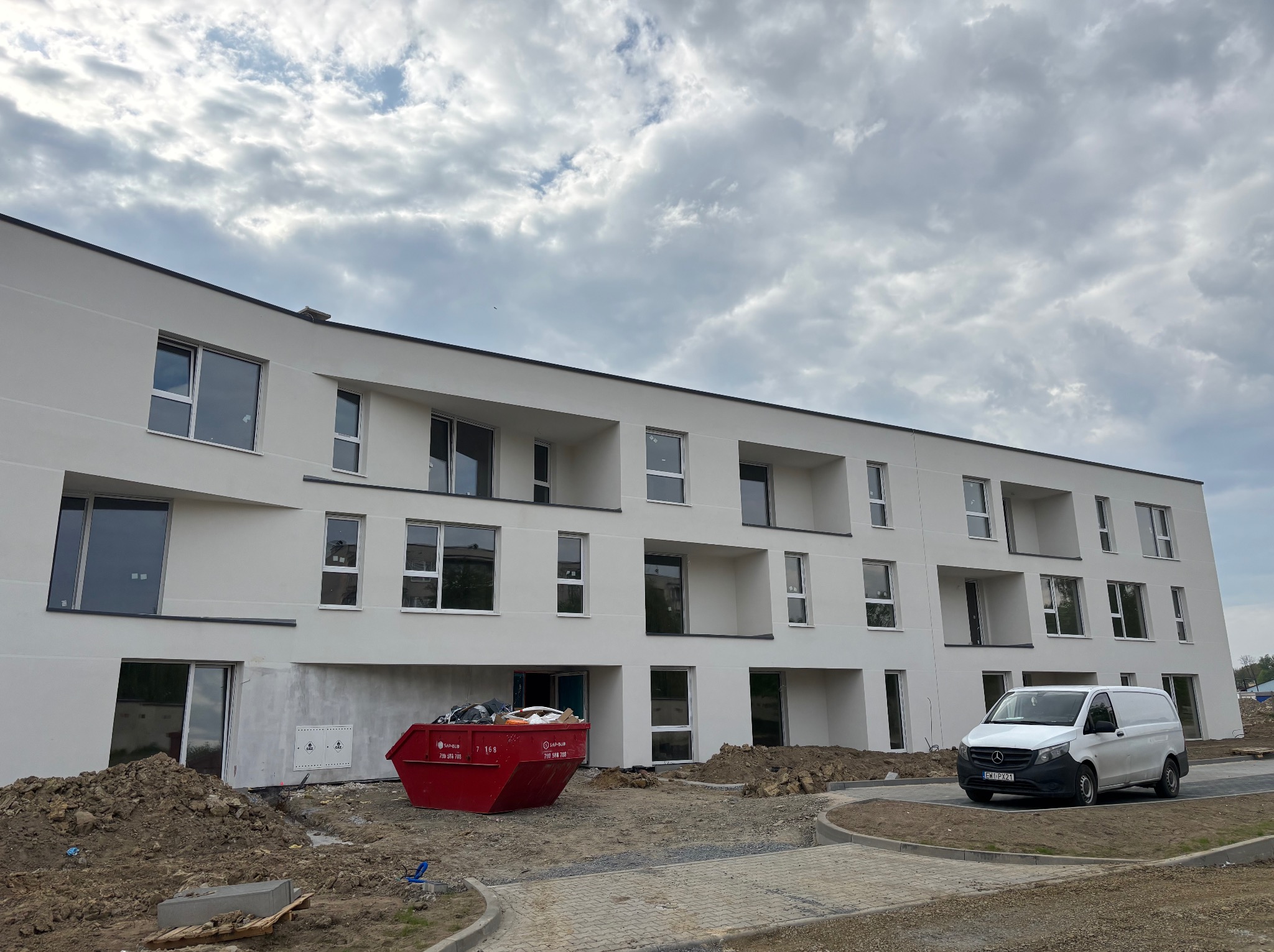 Nowoczesny, biały budynek wielorodzinny w trakcie budowy, z czerwonym kontenerem na gruz i białym vanem dostawczym na pierwszym planie, pochmurne niebo w tle.