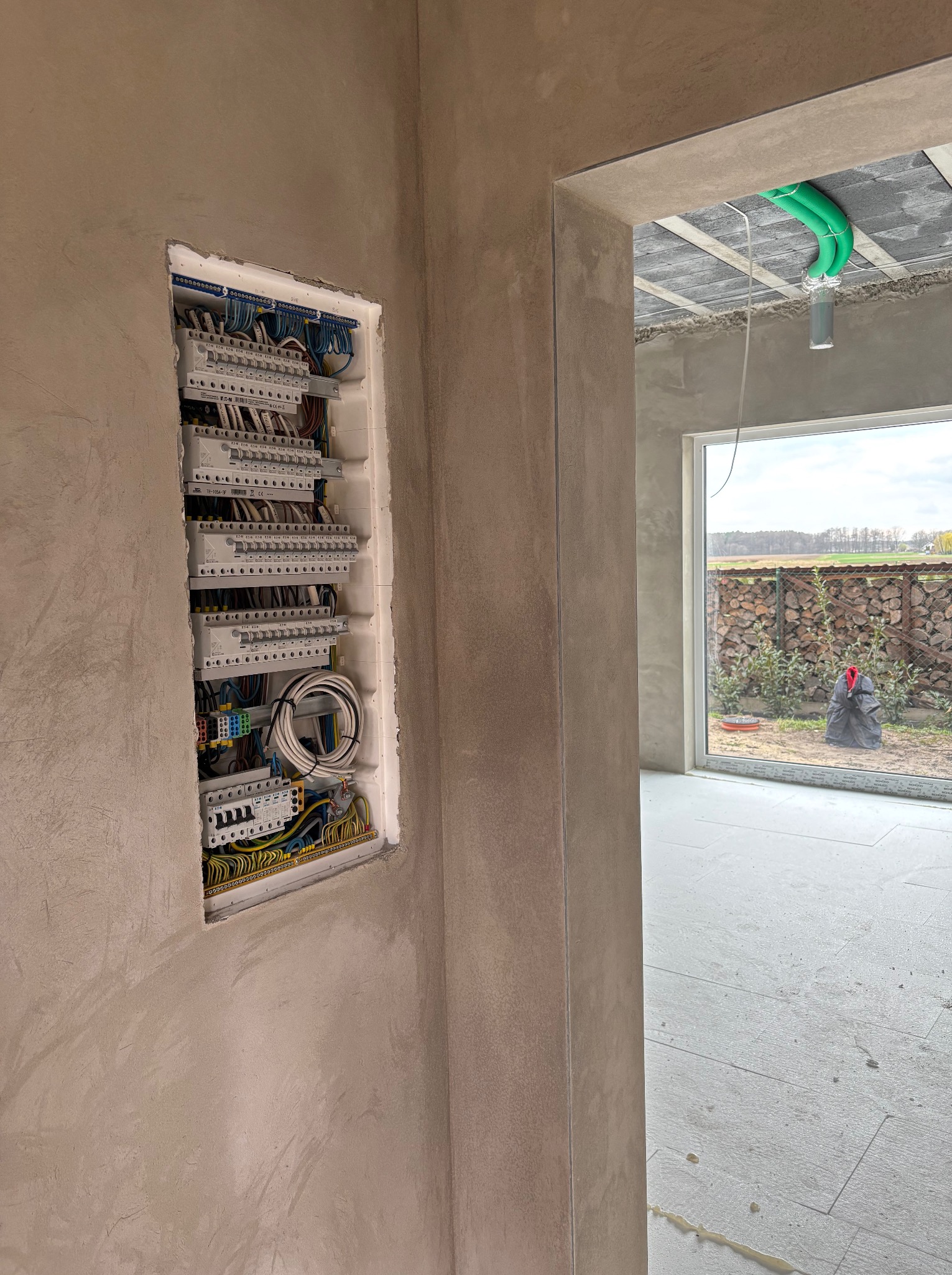 Rozdzielnica elektryczna w surowym wnętrzu domu, widoczne przewody i zabezpieczenia. Przez okno w tle widać ułożone drewno i krajobraz.