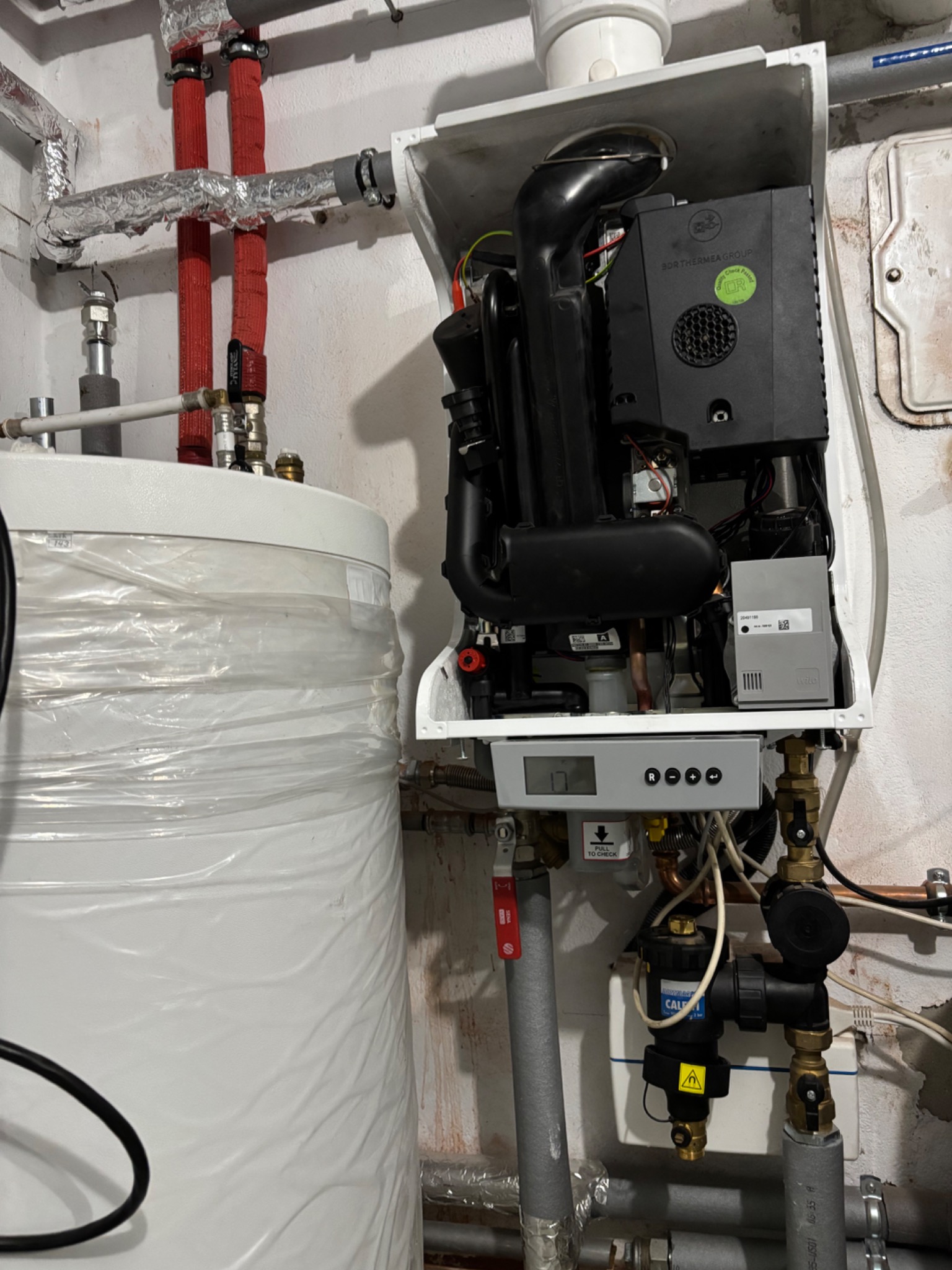 Otwarty piec gazowy z widocznymi elementami instalacji, obok biały zbiornik owinięty folią. Instalacja z czerwonymi rurami w izolacji. Detal techniczny.