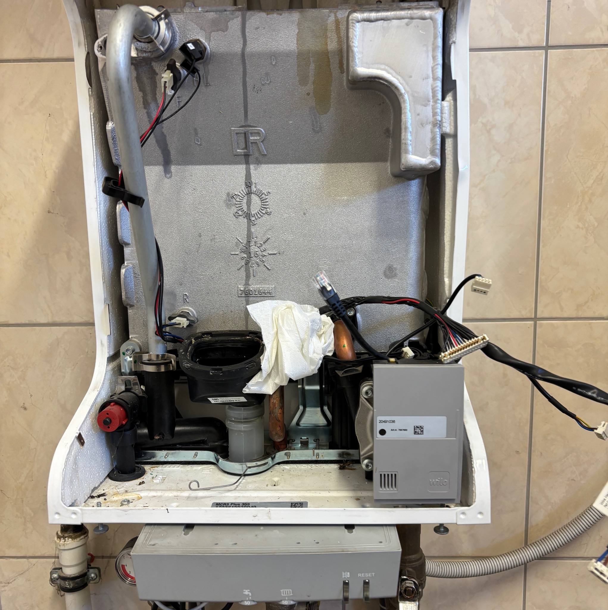 Otwarty piec gazowy z widocznymi elementami instalacji i elektroniką w trakcie przeglądu, z fragmentem białej szmatki wewnątrz i beżowymi płytkami w tle.