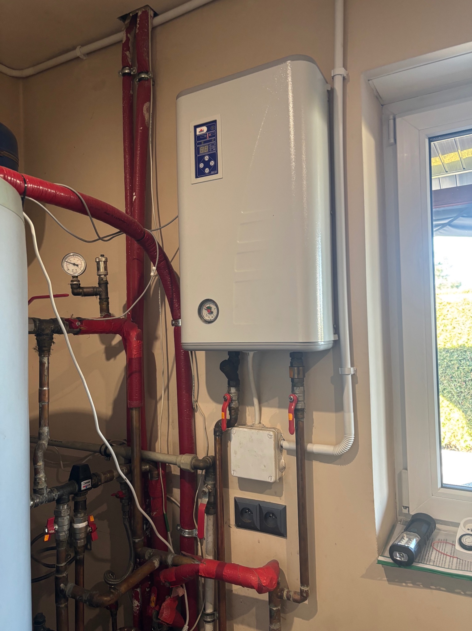 Biały piec gazowy zamontowany na ścianie w Opolu, z widocznymi rurami i instalacją. Okno z widokiem na zewnątrz w tle. Elementy instalacji pomalowane na czerwono.