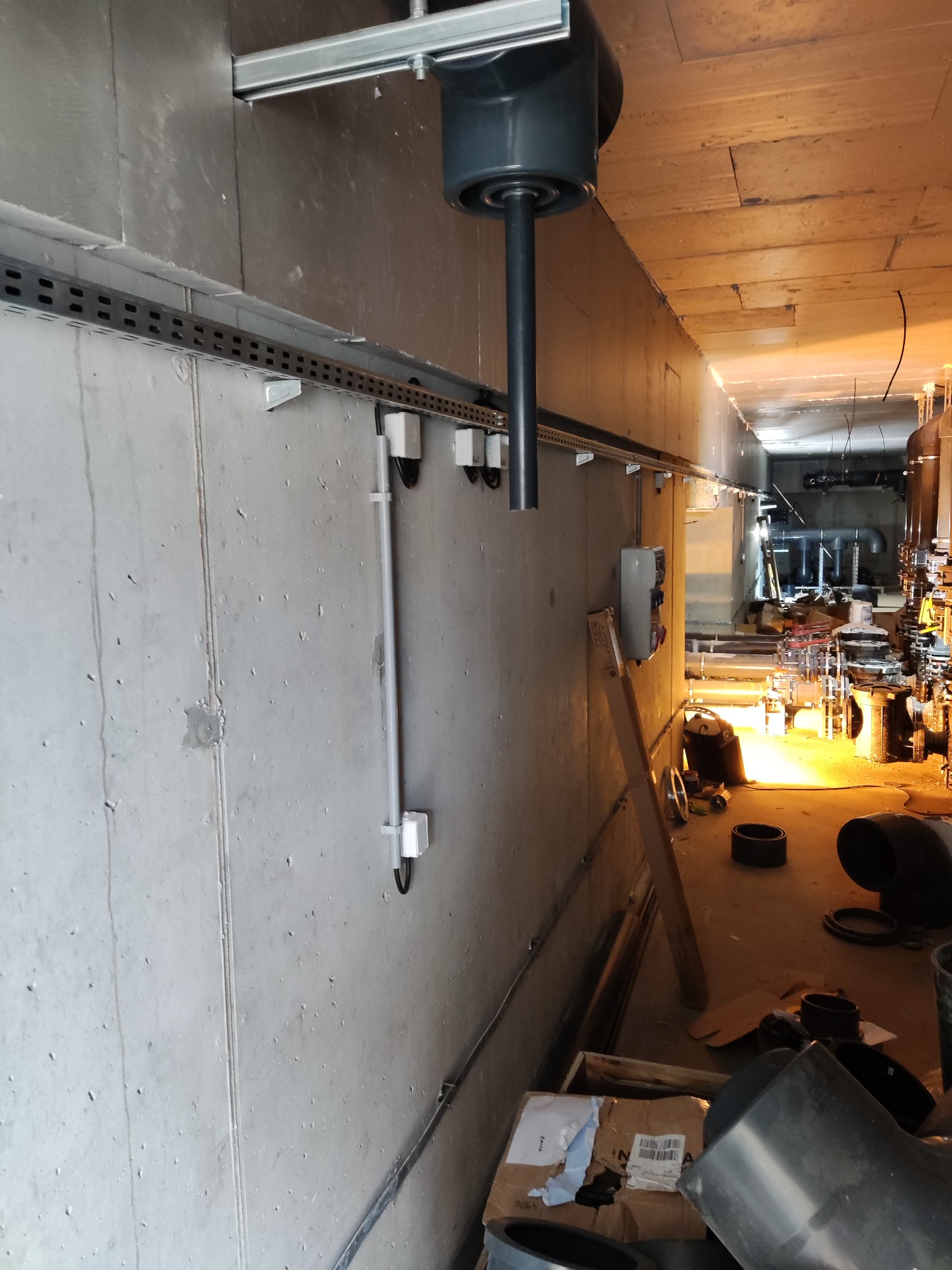 Surowy betonowy korytarz techniczny z widoczną instalacją elektryczną w korytkach, pionową rurą kanalizacyjną, oraz elementami instalacji wodno-kanalizacyjnej w tle.