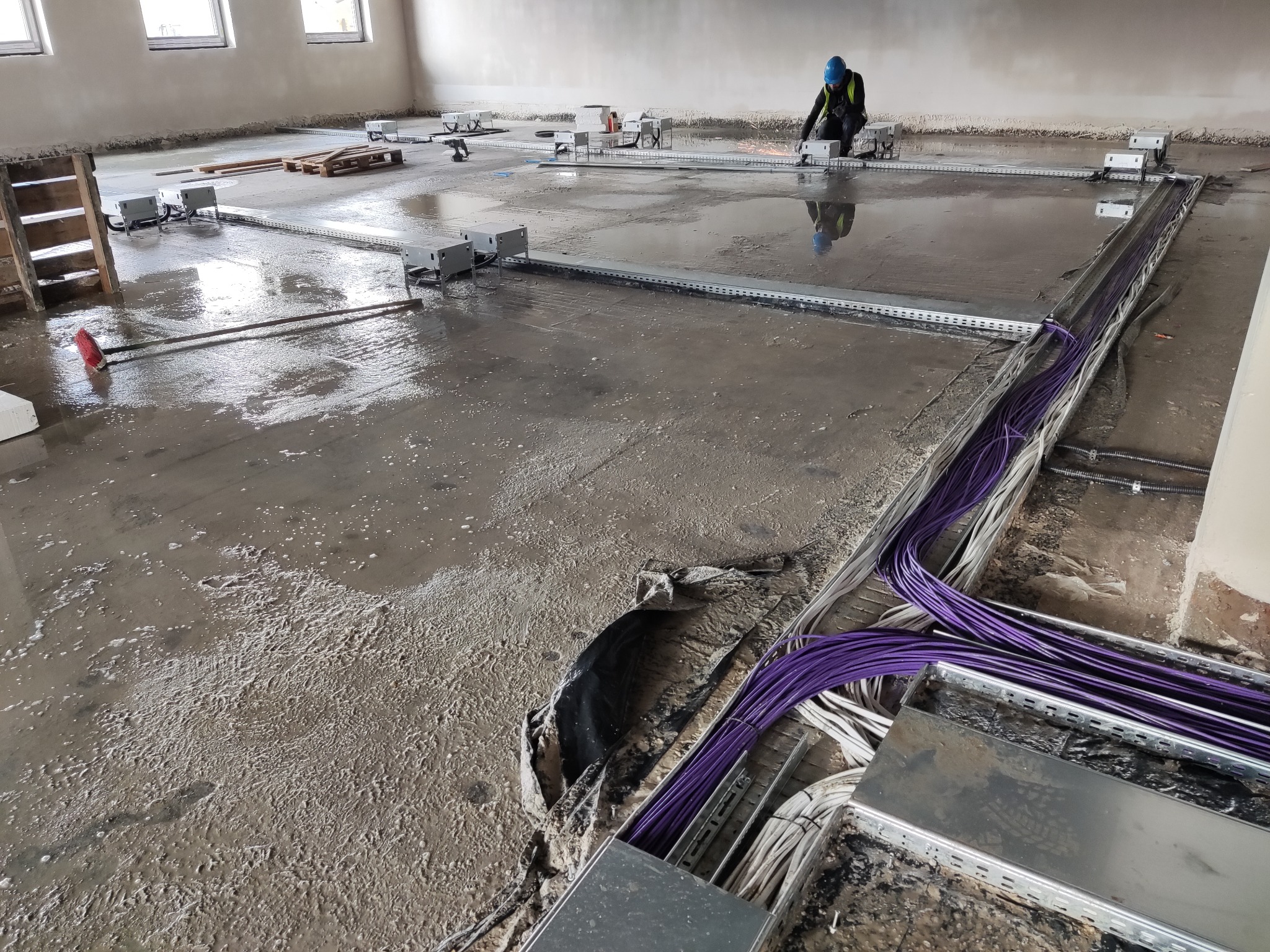 Instalacja okablowania elektrycznego w metalowych korytach na mokrej podłodze podczas budowy, widoczny pracownik w kasku i kamizelce odblaskowej.