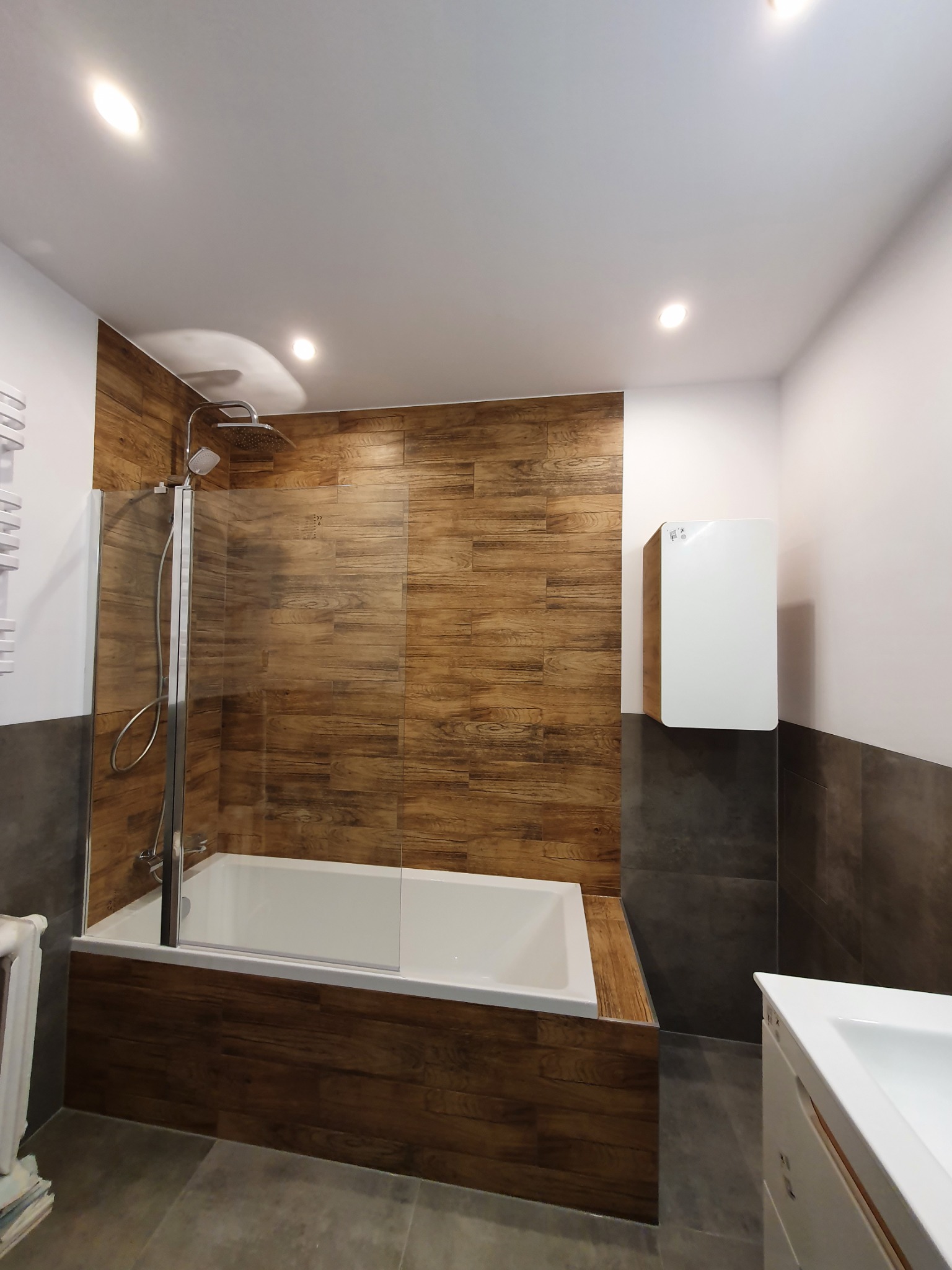 Nowoczesna łazienka z wanną obudowaną panelami imitującymi drewno, przeszklonym parawanem nawannowym i grzejnikiem drabinkowym w stylu retro. Na ścianie biały grzejnik elektryczny, a nad wanną...