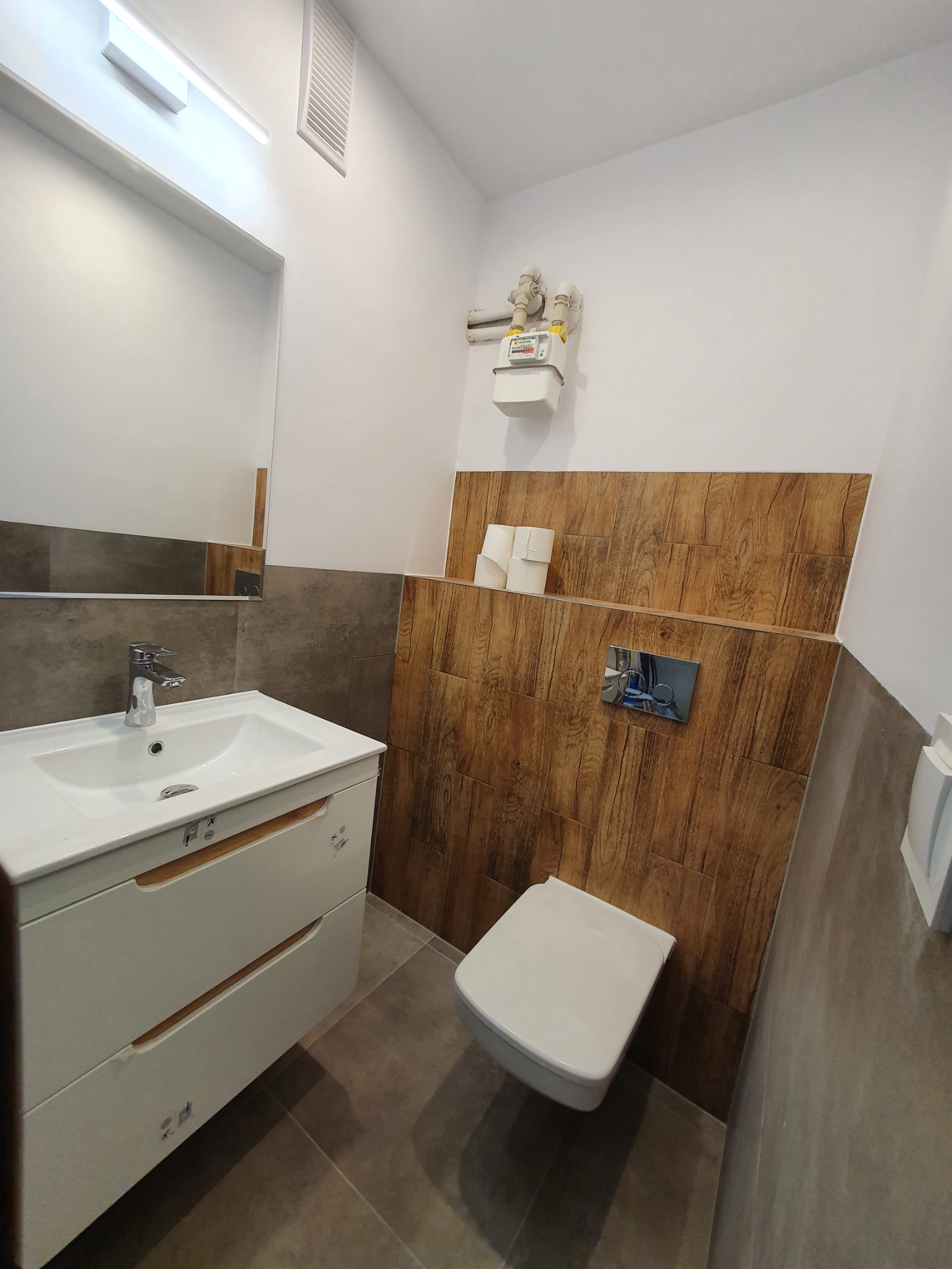 Nowoczesna łazienka po remoncie z umywalką na szafce, toaletą podwieszaną z drewnopodobną zabudową i szarymi płytkami.