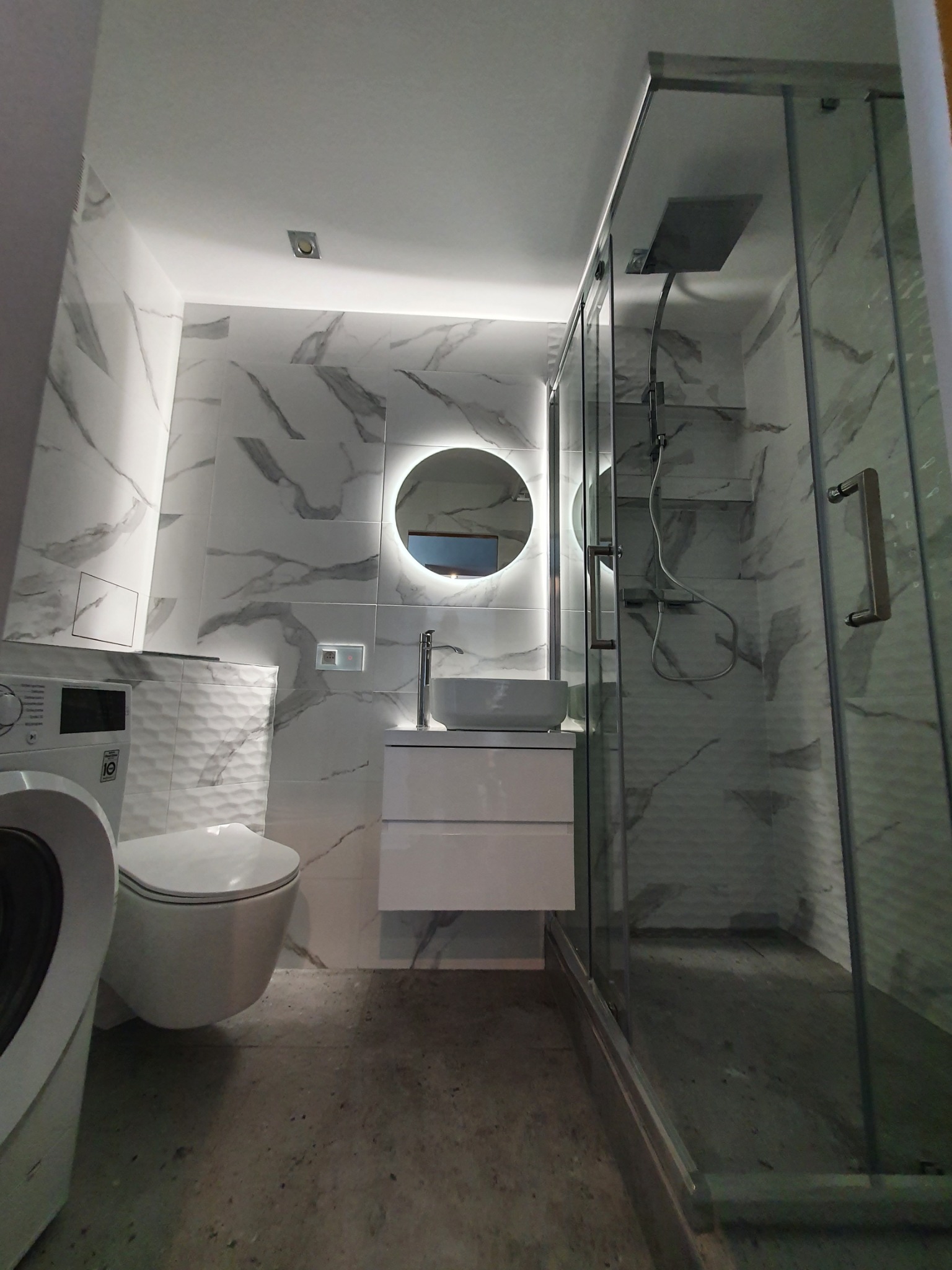 Nowoczesna łazienka z marmurowymi płytkami, okrągłym podświetlanym lustrem, kabiną prysznicową ze szklanymi drzwiami i pralką z przodu.