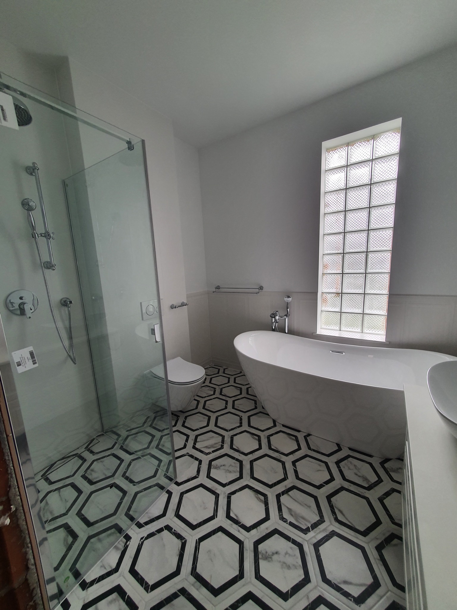 Nowoczesna łazienka z wolnostojącą wanną, kabiną prysznicową ze szklaną ścianką i toaletą podwieszaną, wykończona heksagonalnymi płytkami podłogowymi w kolorze czarno-białym oraz oknem z luksferów.