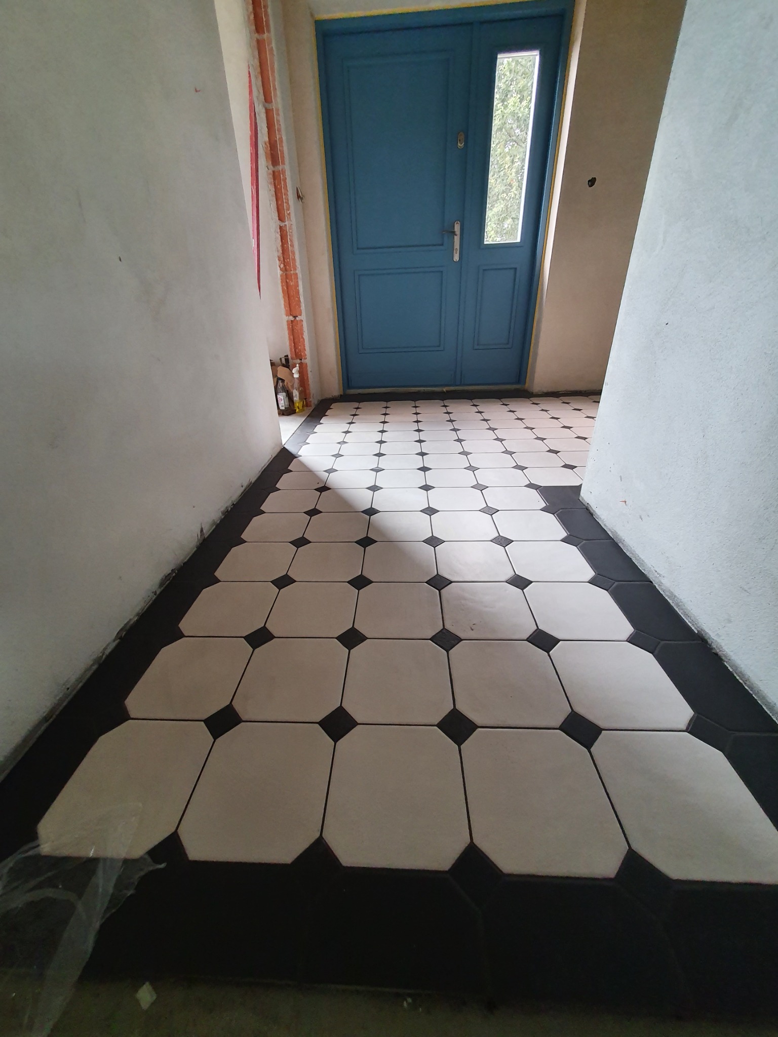 Korytarz w trakcie remontu z nową, czarno-białą podłogą z płytek oktagonalnych i niebieskimi drzwiami wejściowymi; ściany w stanie surowym, widoczne przewody elektryczne.