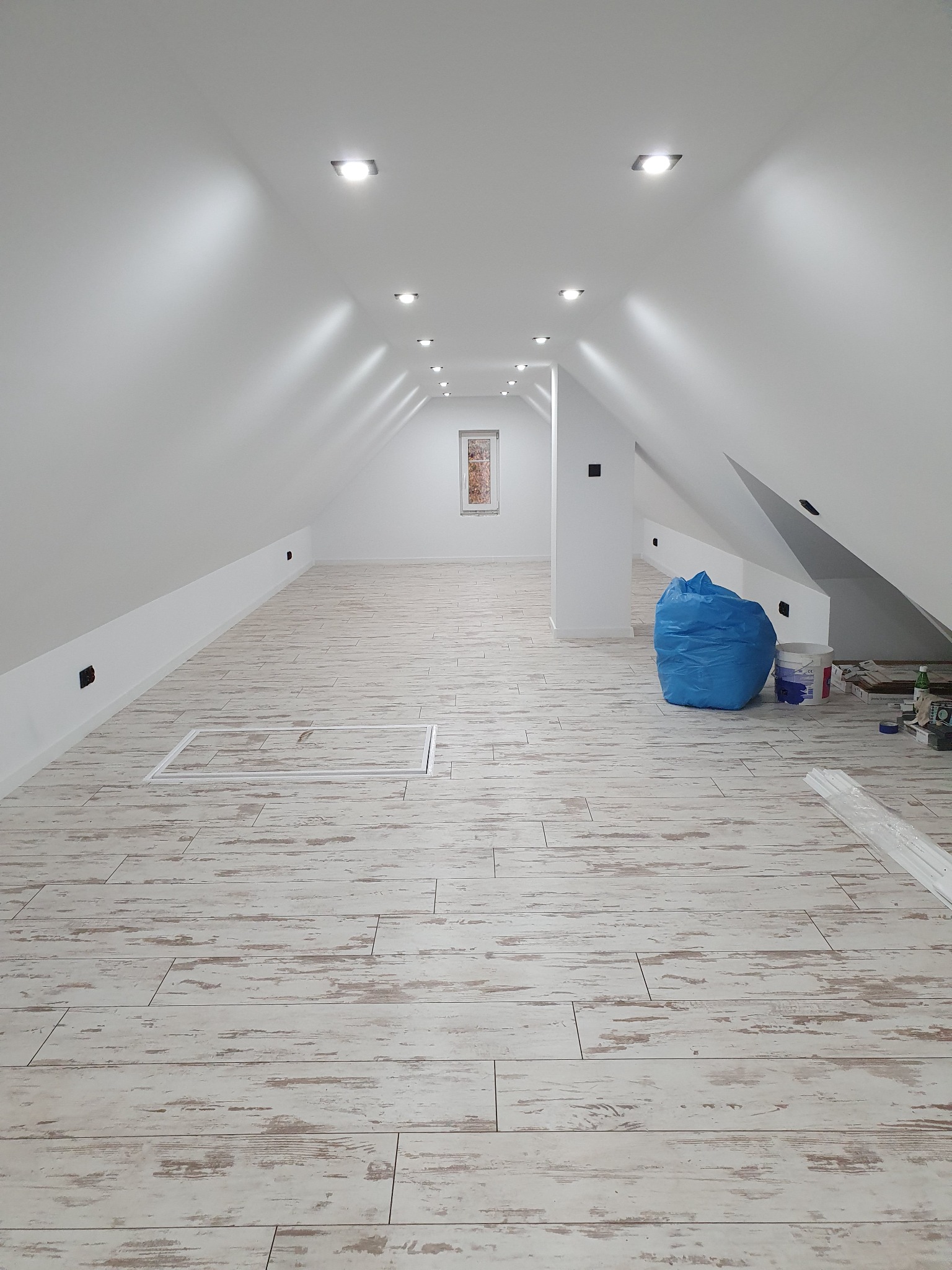 Wykończone poddasze ze skośnymi ścianami, oświetleniem punktowym w suficie i podłogą z paneli imitujących bielone drewno; widoczny niebieski worek z odpadami budowlanymi i akcesoria malarskie.