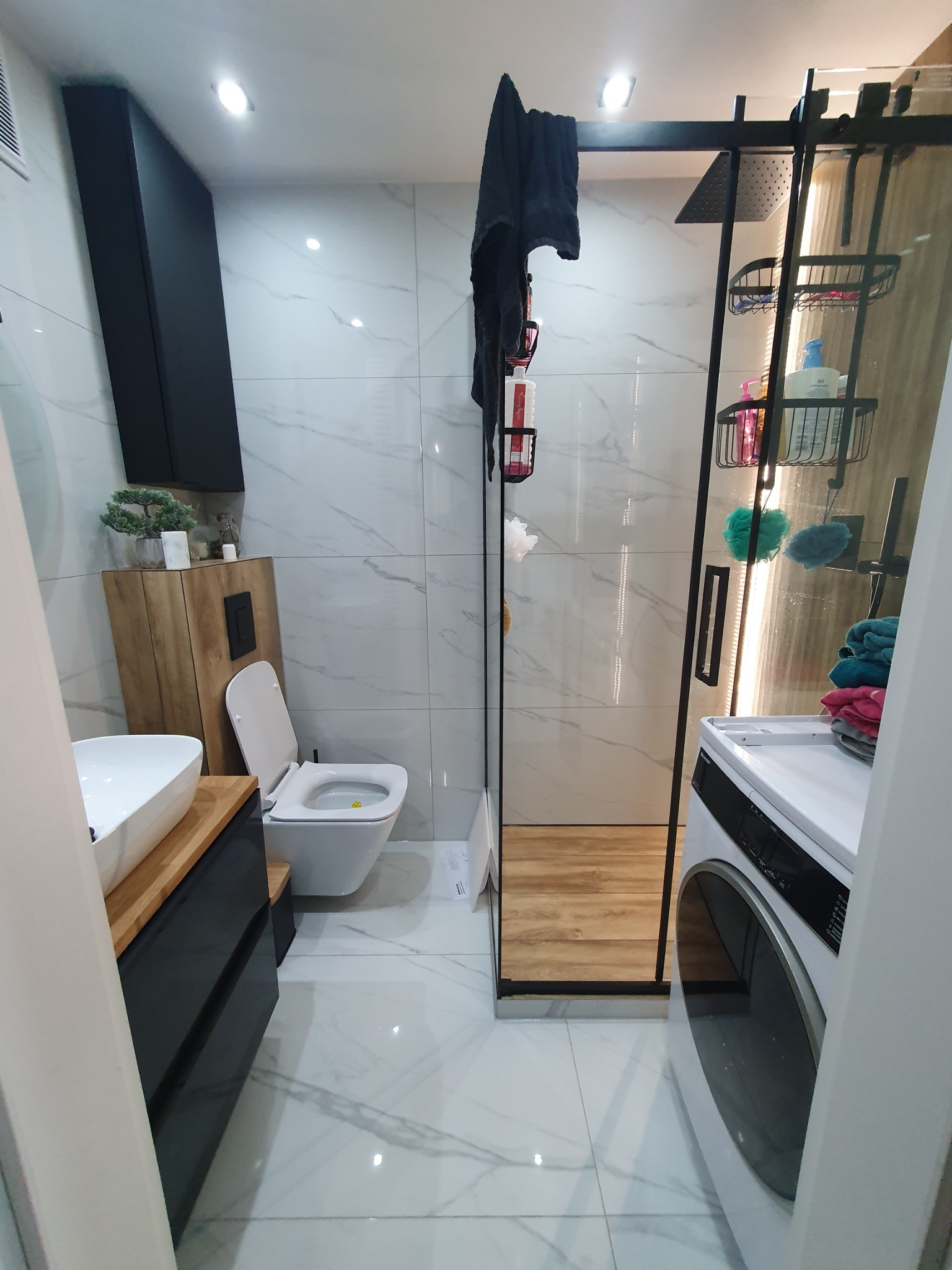 Nowoczesna łazienka z marmurowymi płytkami, kabiną prysznicową typu walk-in z czarną ramą, wiszącą szafką z umywalką na drewnianym blacie, toaletą i pralką.