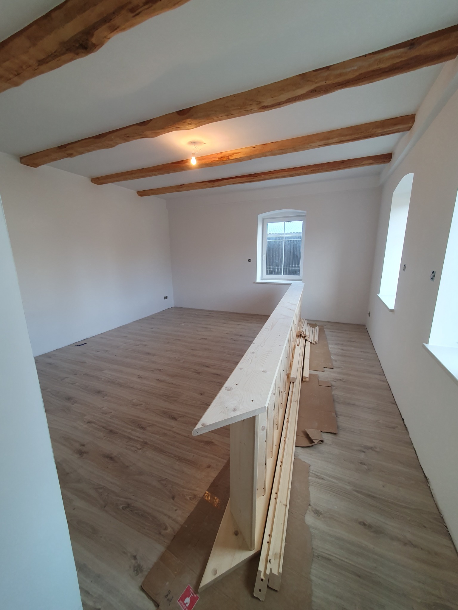 Wnętrze w Bolesławcu z drewnianą podłogą, białymi ścianami i sufitowymi belkami. Częściowo zamontowana balustrada z surowego drewna leży na podłodze.