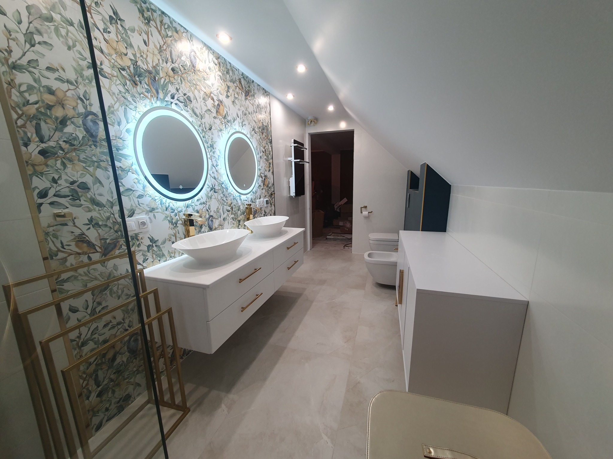 Nowoczesna łazienka na poddaszu z dwoma okrągłymi lustrami z podświetleniem LED, białą szafką z umywalkami nablatowymi, armaturą w kolorze złotym, wzorzystymi płytkami ściennymi z motywem roślinnym...