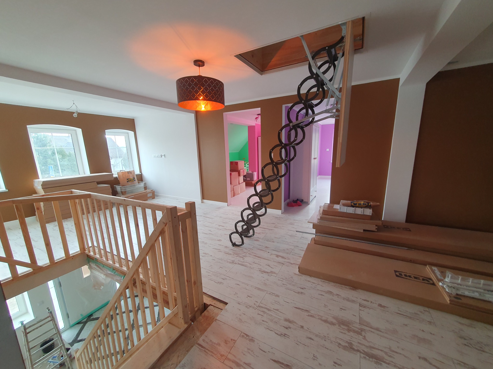 Wnętrze domu w trakcie prac wykończeniowych: rozłożone schody strychowe, drewniane barierki schodowe, podłoga w stylu vintage, kartony z logo IKEA, widok na kolorowe pokoje w tle.