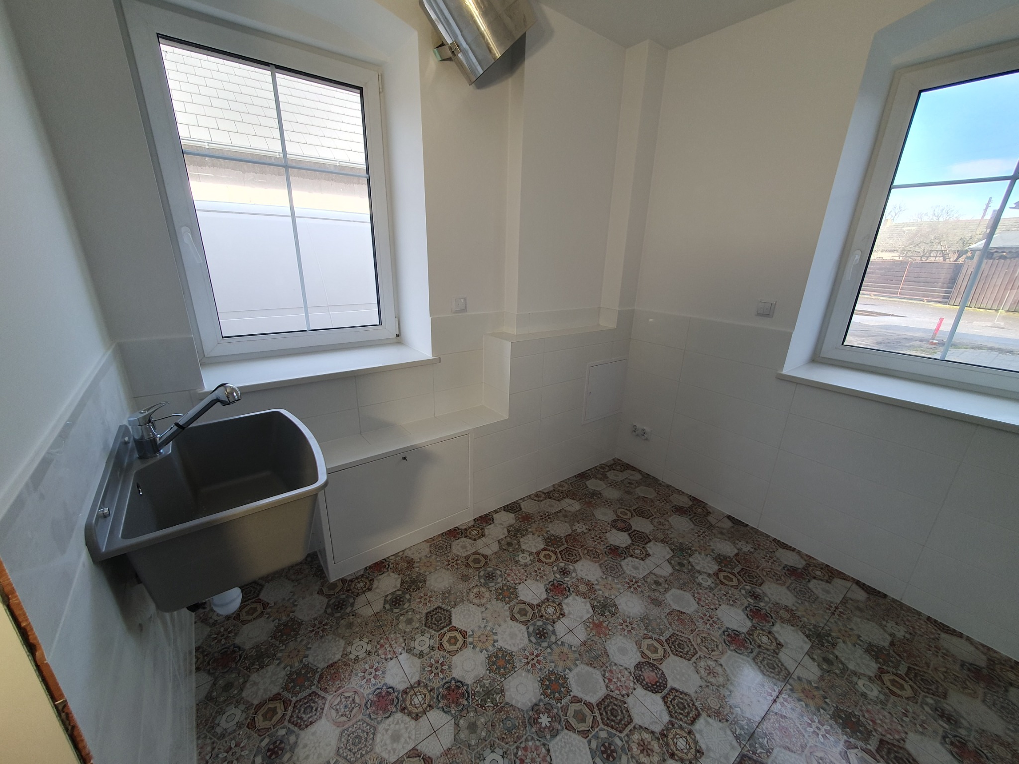 Wnętrze pomieszczenia z umywalką narożną, białe kafelki na ścianach do połowy wysokości, okna z widokiem na zewnątrz, podłoga wyłożona płytkami w stylu patchwork. Metalowa rura wentylacyjna...