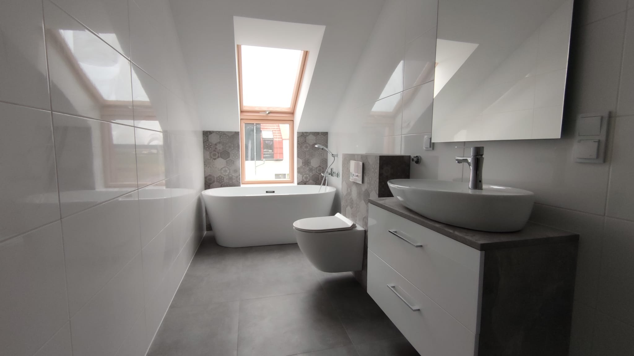 Nowoczesna łazienka na poddaszu z białą wanną wolnostojącą, wiszącą toaletą, umywalką nablatową na szafce z szufladami i oknem dachowym, wykończona białymi płytkami i dekoracyjną mozaiką.
