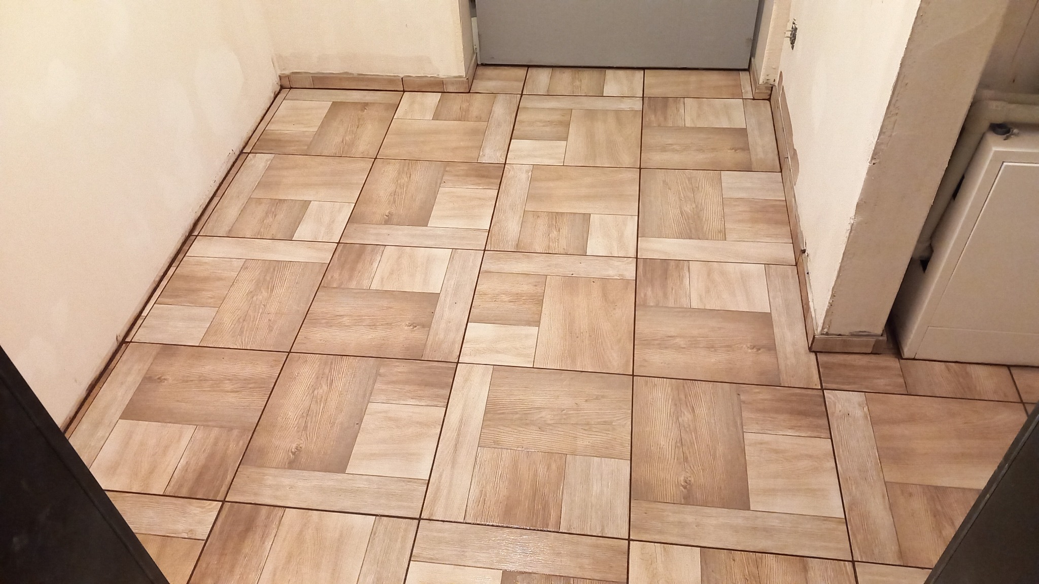 Ułożona podłoga z płytek ceramicznych imitujących drewno w pomieszczeniu z widocznymi fragmentami ścian i futryny drzwi.