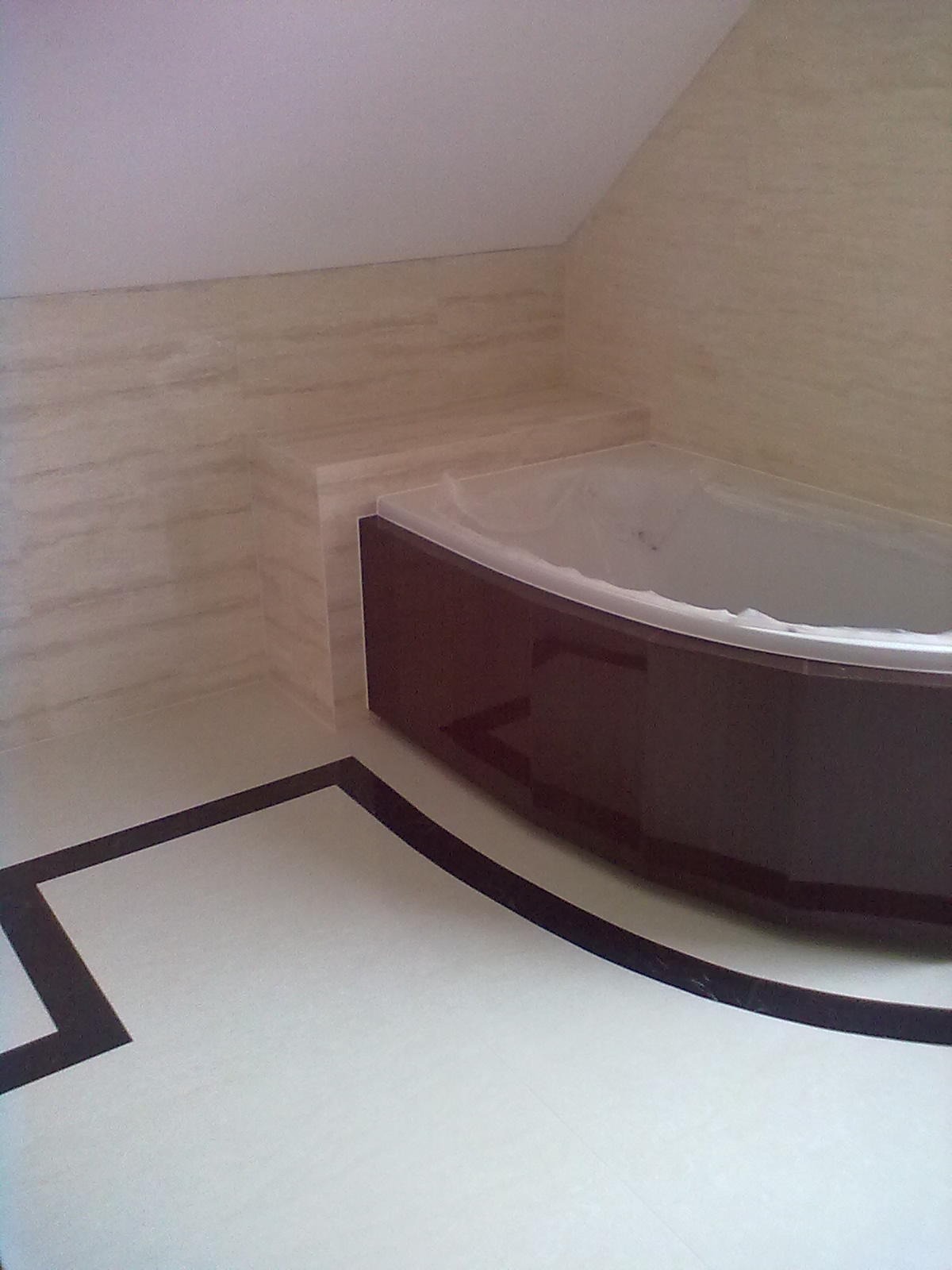 Wykończona łazienka w jasnych odcieniach beżu i bieli z ciemnobrązową obudową wanny narożnej i geometrycznym wzorem z ciemnych płytek na podłodze.