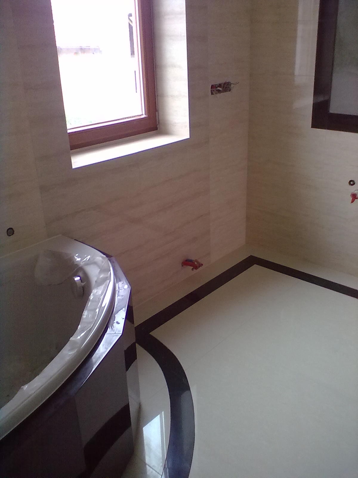 Wykończona łazienka z beżowymi płytkami imitującymi marmur, ciemnobrązową obwódką na podłodze i wanną z hydromasażem, widok na okno i wystające przewody elektryczne.