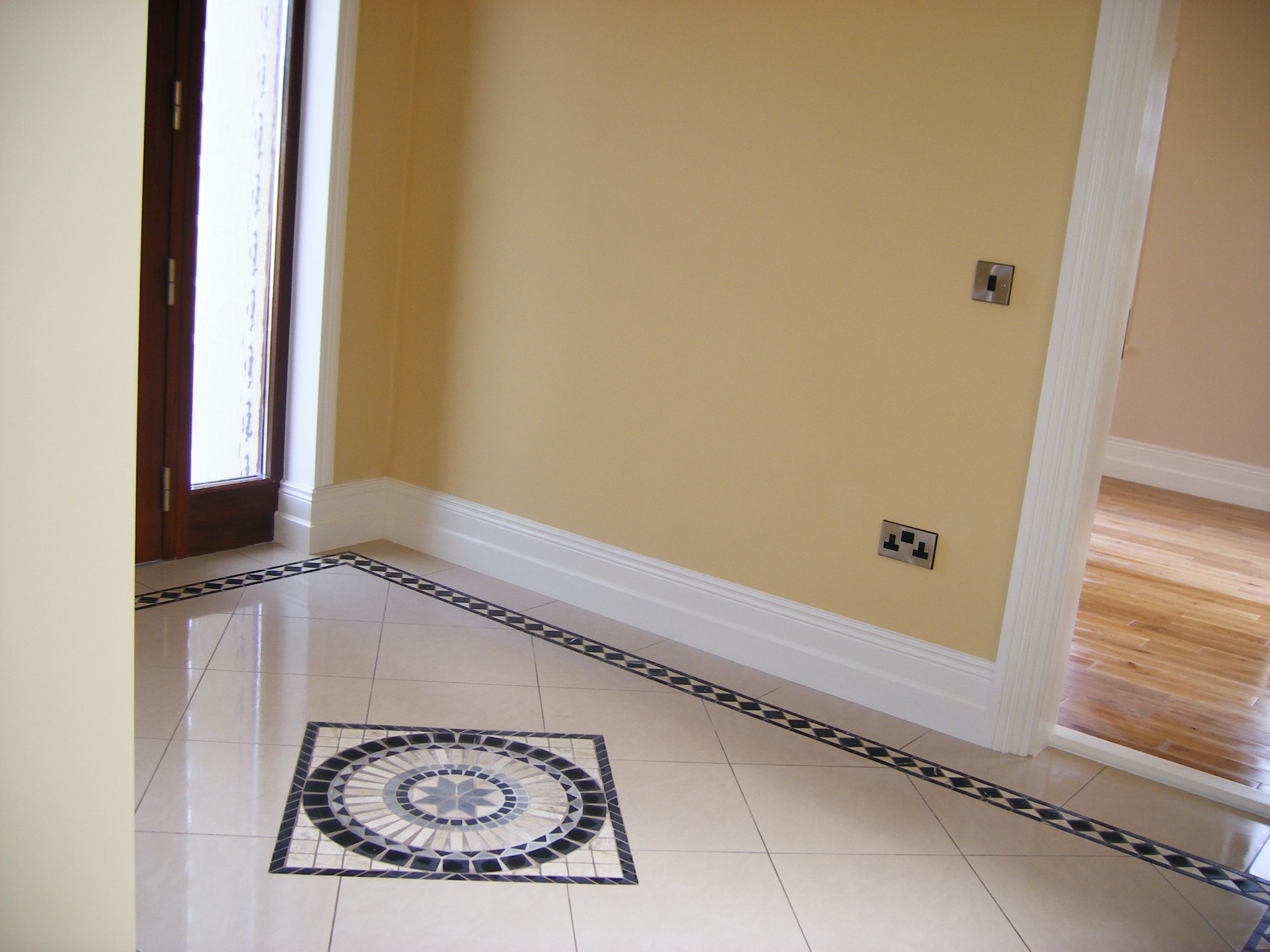 Jasne, błyszczące płytki podłogowe z dekoracyjnym wzorem mozaiki w kształcie rozety i bordiury, widoczny fragment wejścia z drewnianymi drzwiami i gniazdka elektryczne na żółtej ścianie.