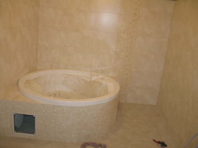 Beżowa łazienka z okrągłą wanną obudowaną mozaiką, płytki ścienne z delikatnym wzorem, narzędzia na podłodze.