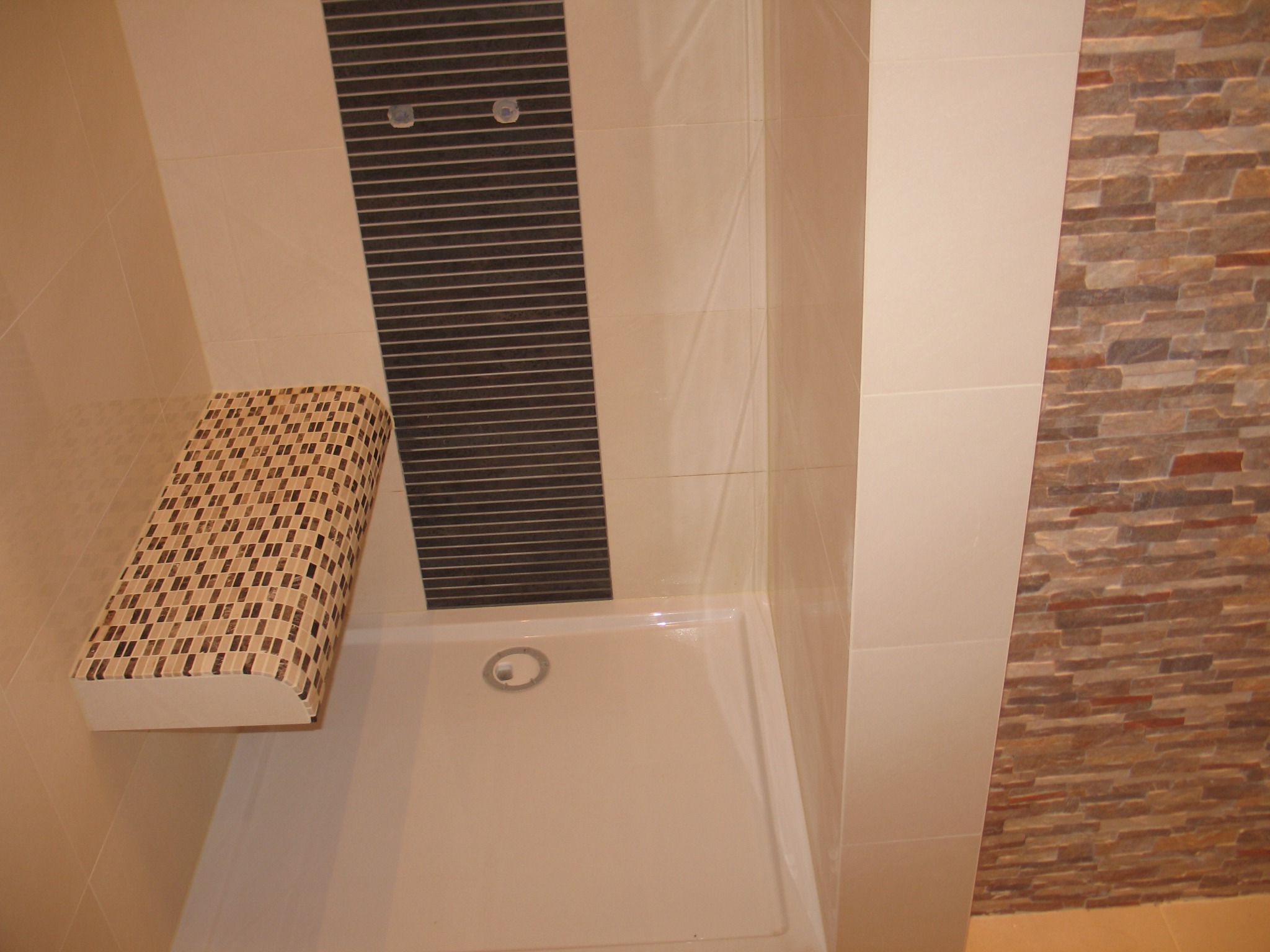 Wykończona kabina prysznicowa z białą brodzikiem, półką wykończoną mozaiką oraz ścianami z płytek w odcieniach beżu i brązu, z dekoracyjnym panelem z poziomych pasków.