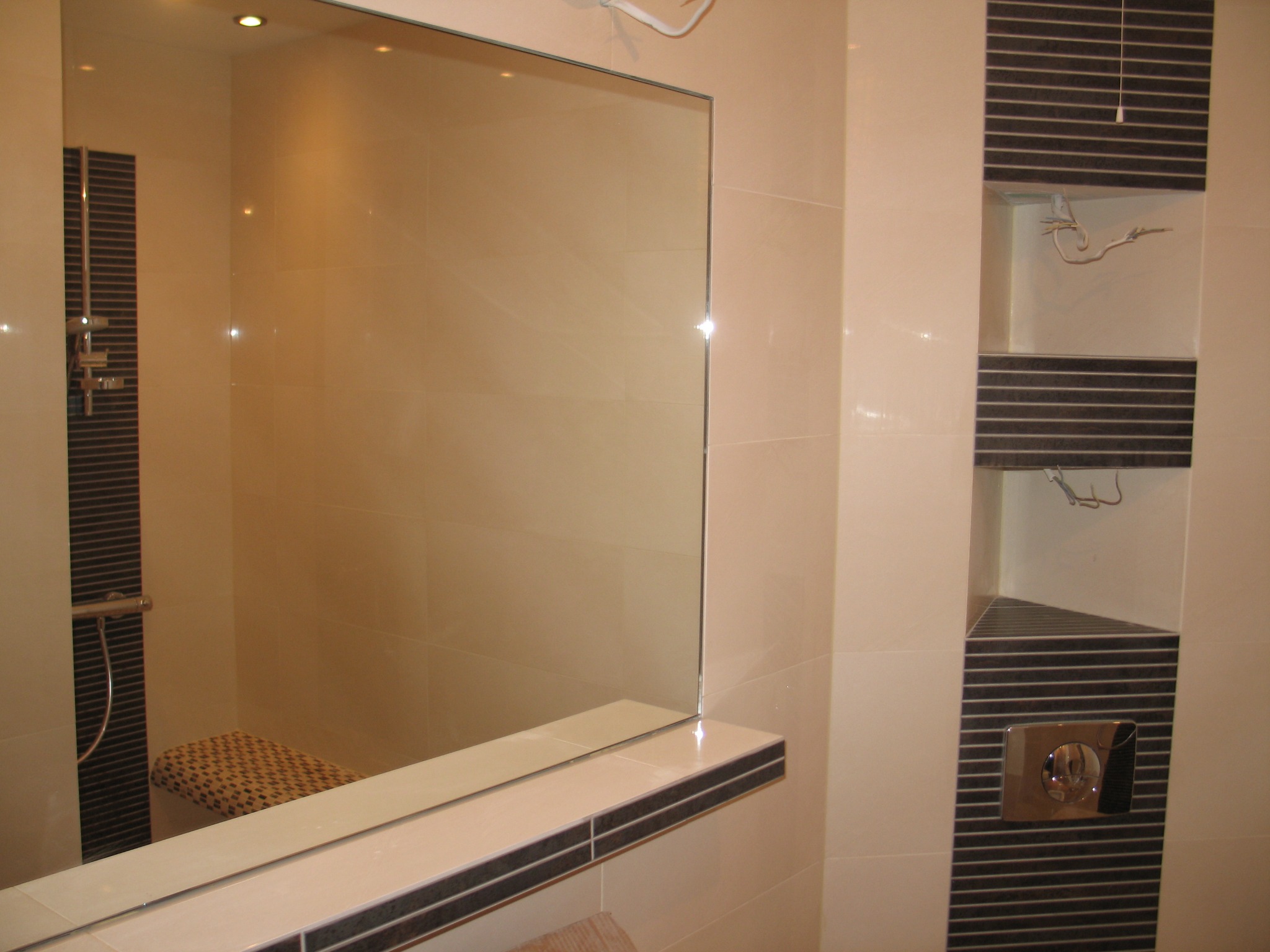 Wykończona łazienka z beżowymi płytkami, dużym lustrem z półką i wnęką z dekoracyjnymi elementami oraz panelem z przyciskiem spłuczki.