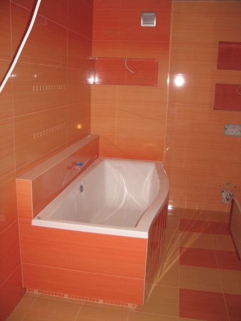 Pomarańczowa łazienka z wanną obłożoną płytkami, widoczne płytki na ścianach i podłodze, wentylator w górnej części ściany.