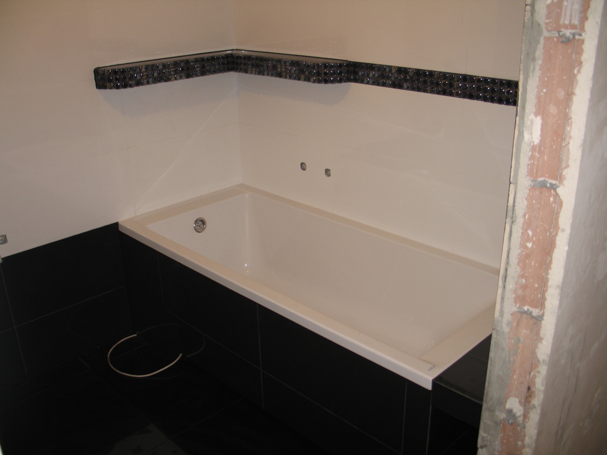 Nowoczesna łazienka z wanną obłożoną białymi płytkami, czarne płytki na podłodze i dolnej części ściany, dekoracyjny pasek z ciemnych kulek pod sufitem, widoczny fragment surowej ściany w tle.